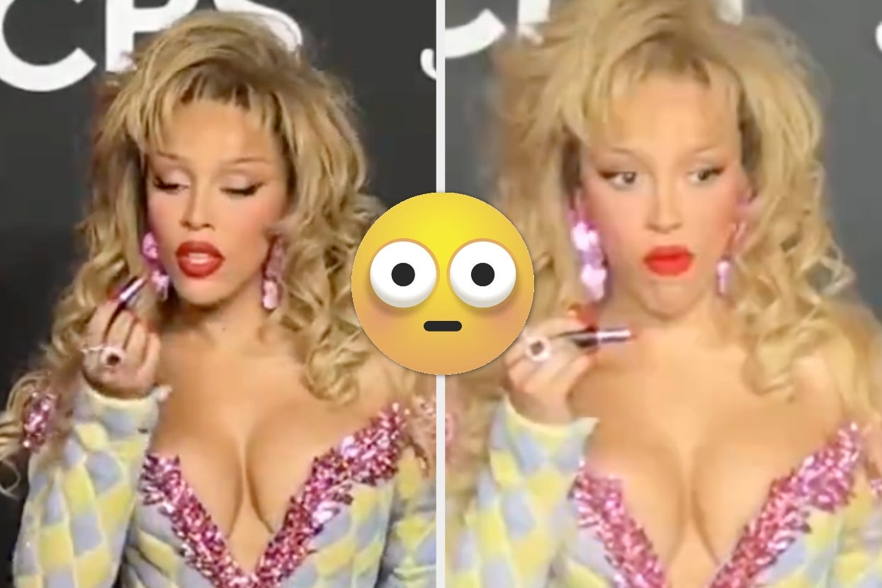 Person on red carpet wearing a deep-V outfit with glitter accents, applying lipstick with a focused expression