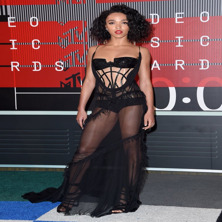 A person poses on a red carpet wearing a sheer black gown with a fitted bodice and high-slit skirt
