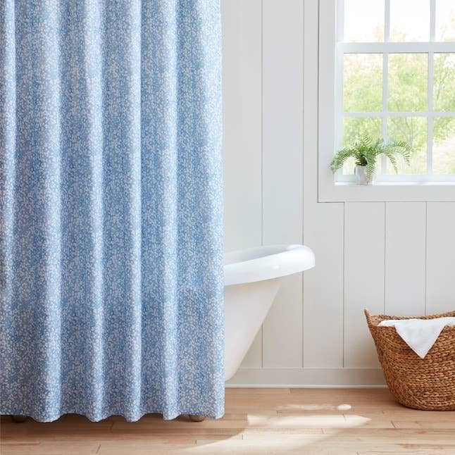 Bathtub with a floral-patterned ablution curtain adjacent to a window, white-paneled walls, and a wicker basket