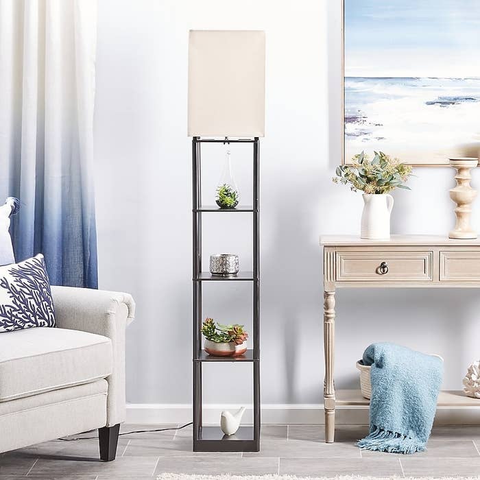 Tall level lamp with quadrate shade, featuring shelves holding tiny plants and decor. Near a sofa and console array successful a coastal-themed room
