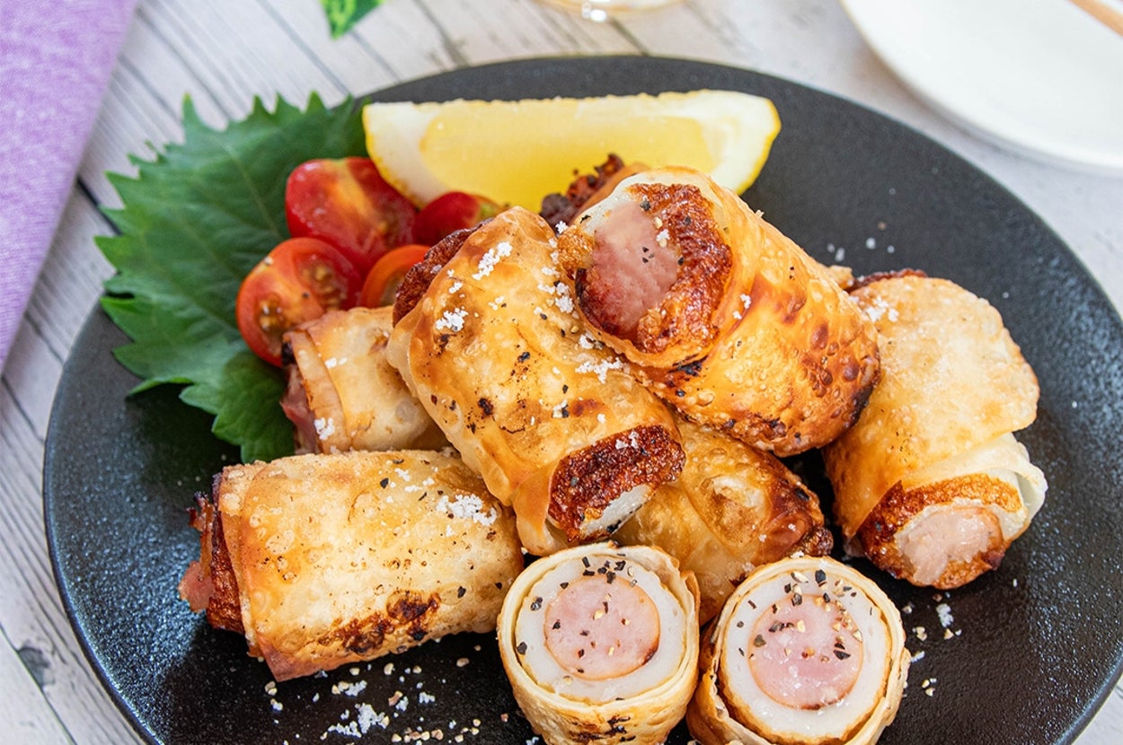 ソーセージがちくわで包まれた料理が盛り付けられた黒い皿。添えられたトマトとレモンが鮮やか。