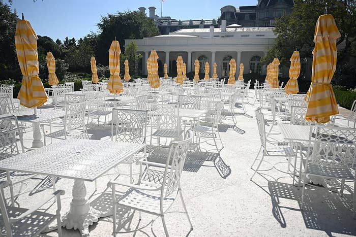 Outdoor patio with empty white metal tables and chairs, surrounded by yellow striped umbrellas. A building with columns is in the background