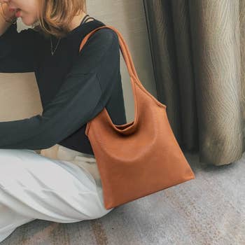 Person seated adjacent a curtain, wearing a achromatic apical and achromatic pants, showcasing a stylish brownish leather enarthrosis bag
