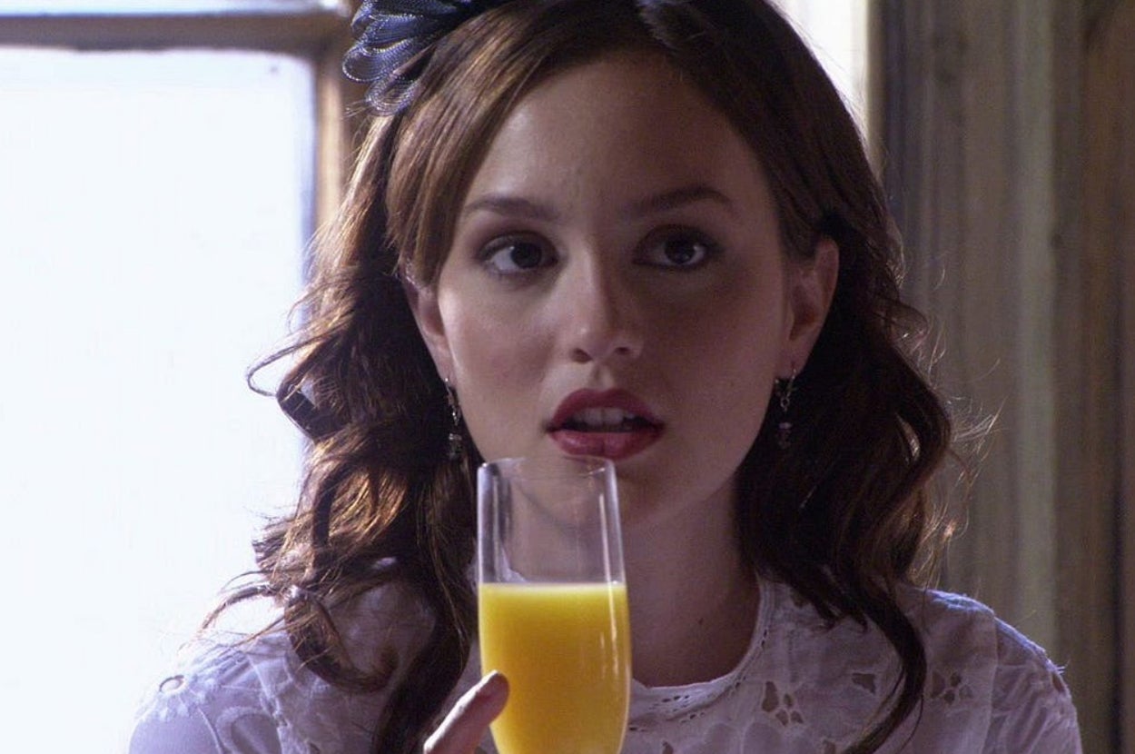 Person with long hair holds a glass of orange juice, wearing a lace top and earrings, looking thoughtful