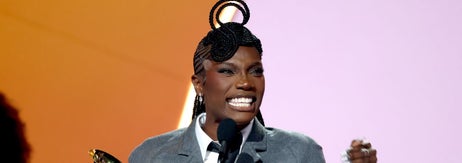 Person holding a Grammy award, speaking at a microphone, wearing a tailored outfit with a unique hairstyle on stage
