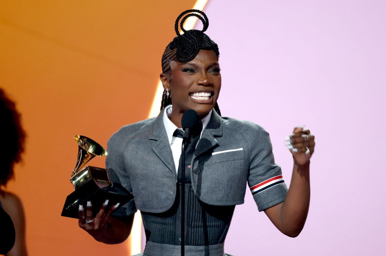 Person holding a Grammy award, speaking at a microphone, wearing a tailored outfit with a unique hairstyle on stage