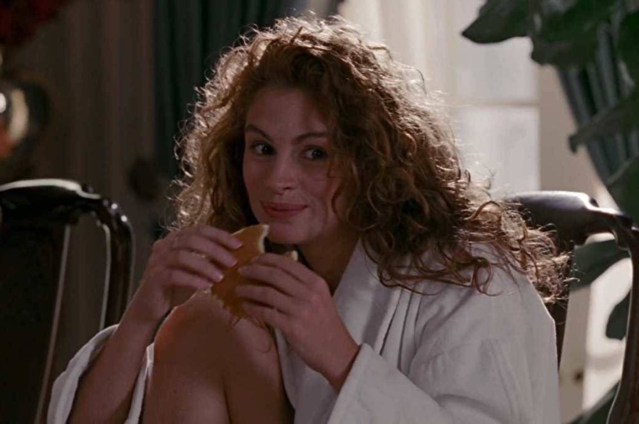 A person with curly hair, wearing a robe, is seated and smiling while holding a sandwich indoors