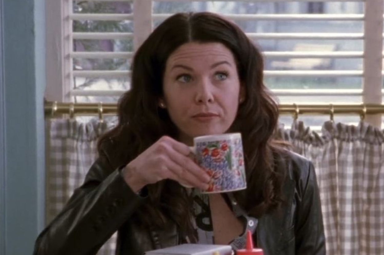 Person with long hair sitting at a table, holding a floral mug and looking into the distance with a thoughtful expression