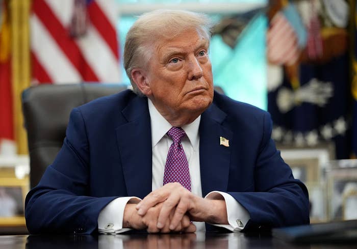 A person in a suit and tie sits at a desk, looking attentive with hands clasped. Flags and framed photos are visible in the background