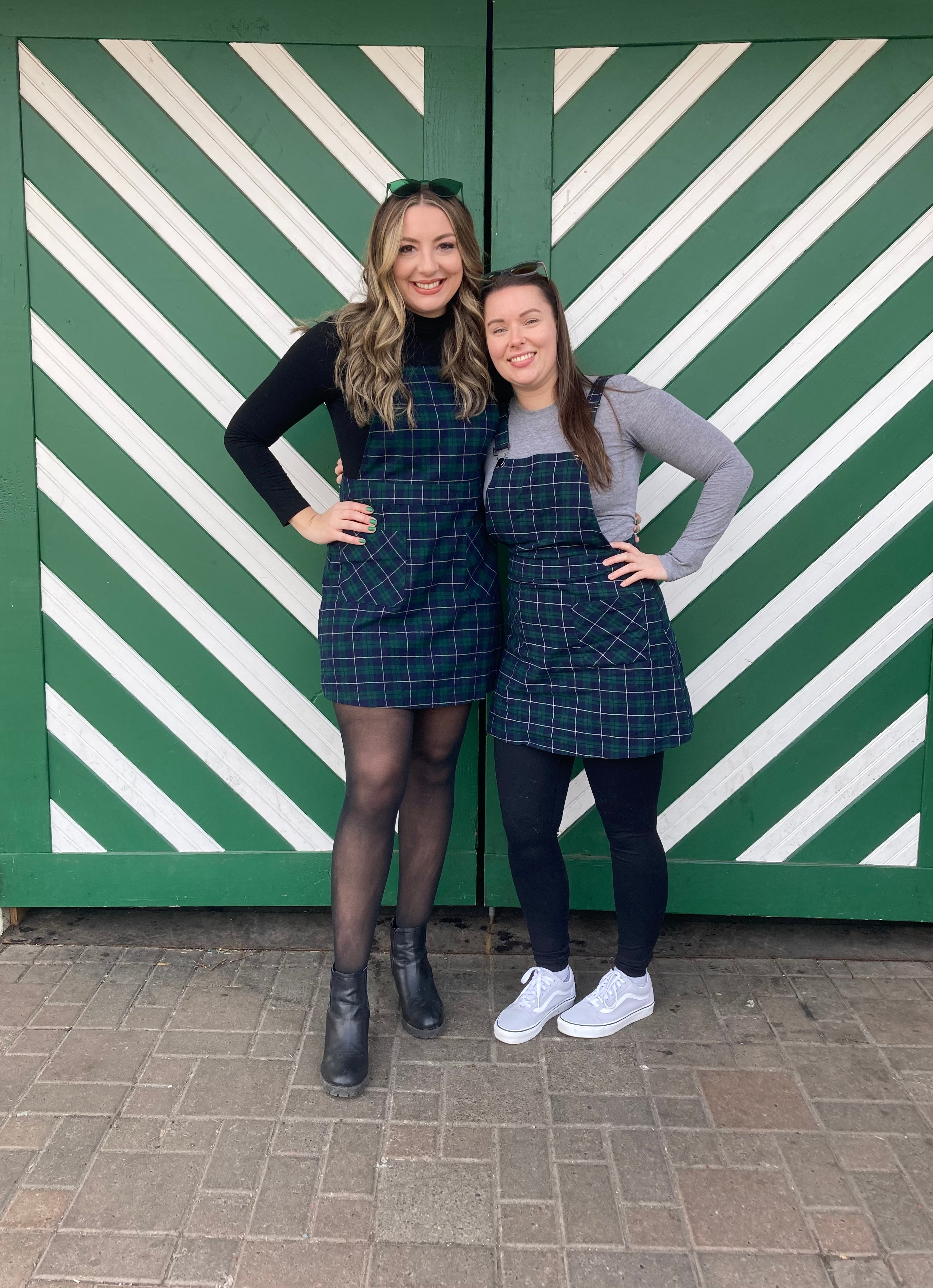 Two radical   smiling, wearing plaid dresses implicit    tops, 1  successful  boots and the different   successful  sneakers, lasting  successful  beforehand   of a greenish  striped background