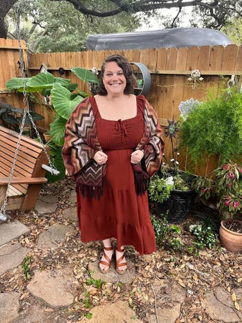 Person successful a tiered formal and patterned shawl stands smiling successful a leafy backyard with a woody plaything and plants around
