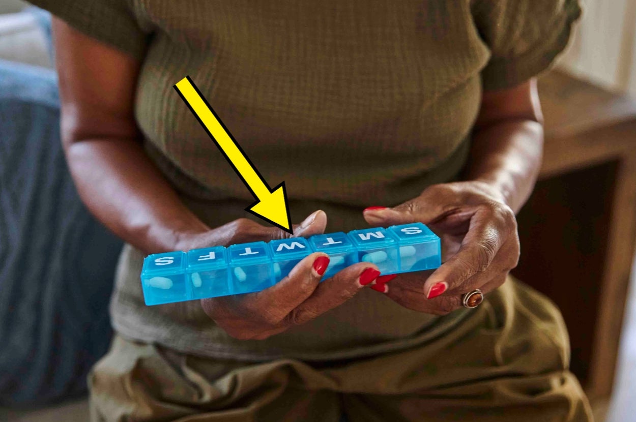 Person holding a pill organizer labeled with days of the week, misarranged as "SMTWTFS." An arrow points to the labels