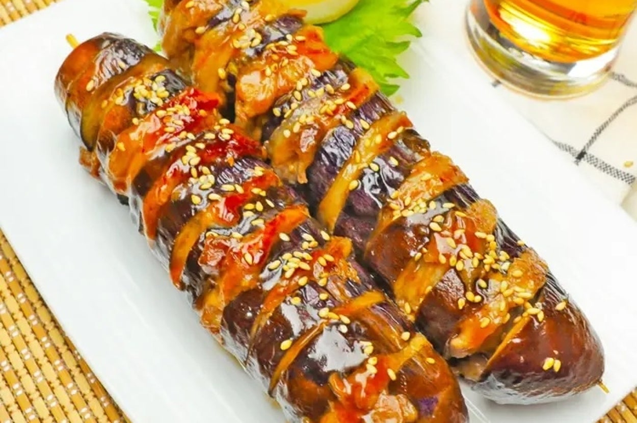 焼き物のナスが串に刺さり、ゴマとソースがかかっている。背景にグラスが見える。
