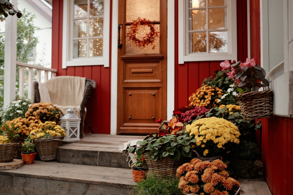 Cozy porch with wooden steps, decorated with baskets of fall flowers, a woven chair, and a lantern by the door adorned with a fall wreath