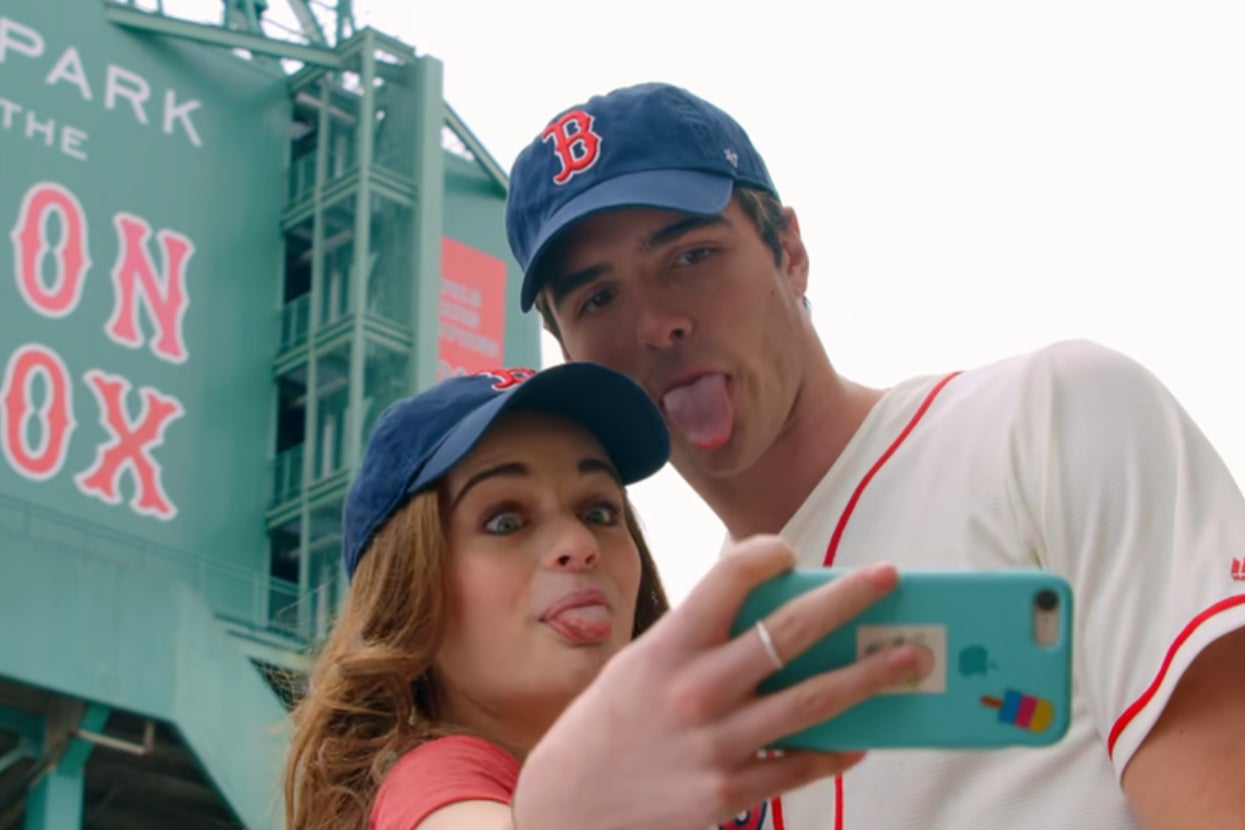 Joey King and Jacob Elordi in "The Kissing Booth" wearing Boston Red Sox baseball caps, taking a selfie outside Fenway Park. They have playful expressions, sticking their tongues out