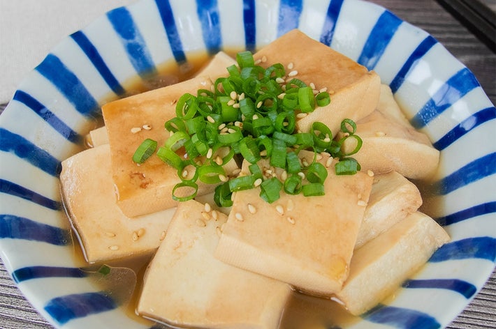 皿に盛られた豆腐料理。スライスし加熱された豆腐に小ネギと白いりごまがトッピングされ、たれがかかっている。