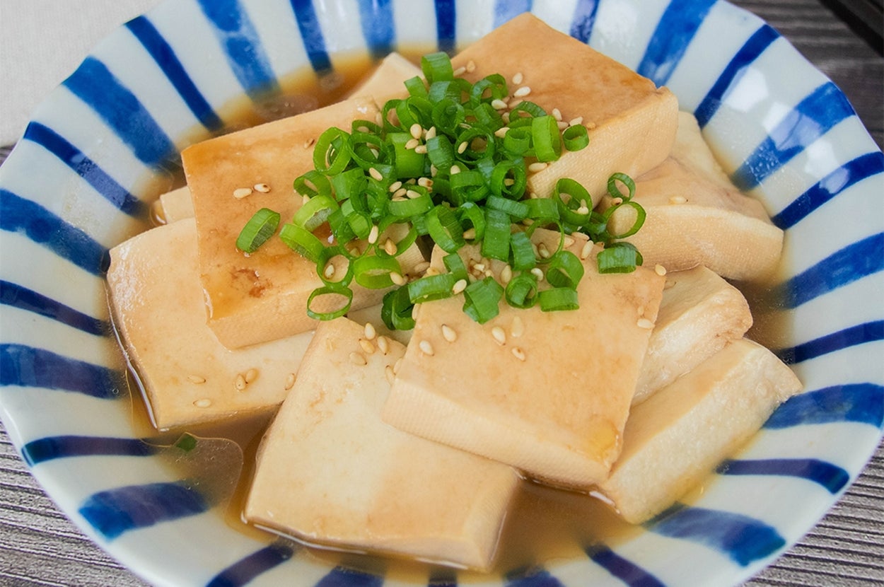 皿に盛られた豆腐料理。スライスし加熱された豆腐に小ネギと白いりごまがトッピングされ、たれがかかっている。