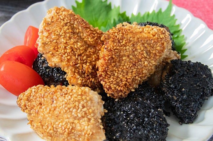 皿の上にゴマをまぶしたチキン、黒ゴマをまぶした揚げ物、プチトマトが盛られている。背景にピンクの布が見える。