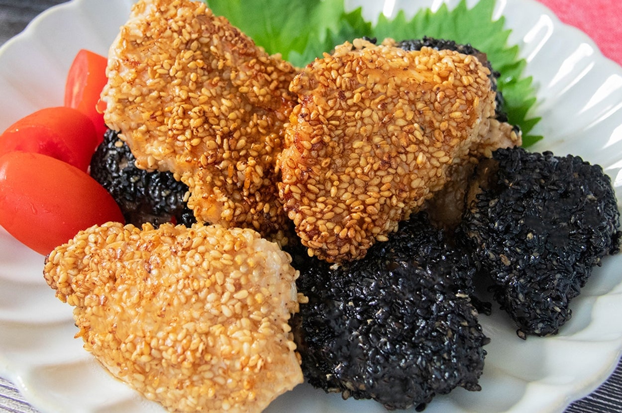 皿の上にゴマをまぶしたチキン、黒ゴマをまぶした揚げ物、プチトマトが盛られている。背景にピンクの布が見える。