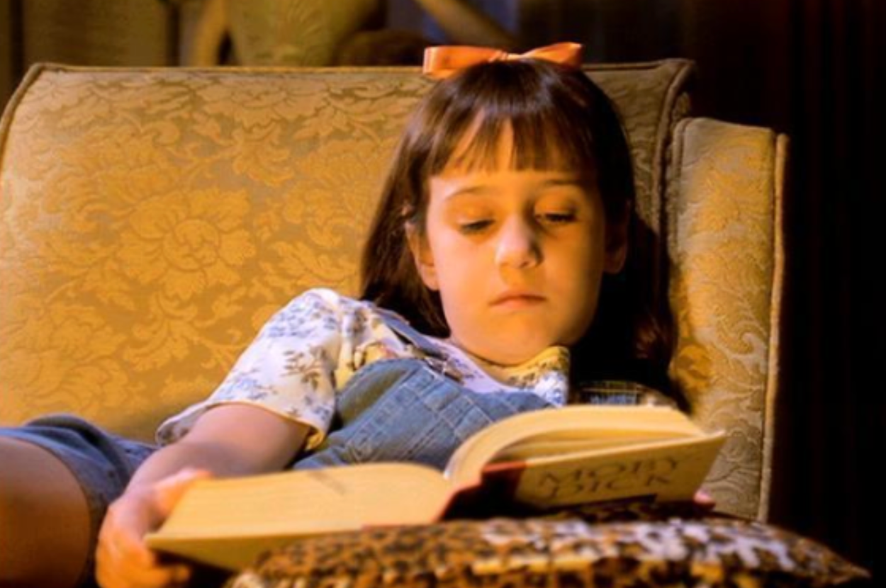 A young girl with a bow in her hair reads a book while relaxing on a patterned armchair