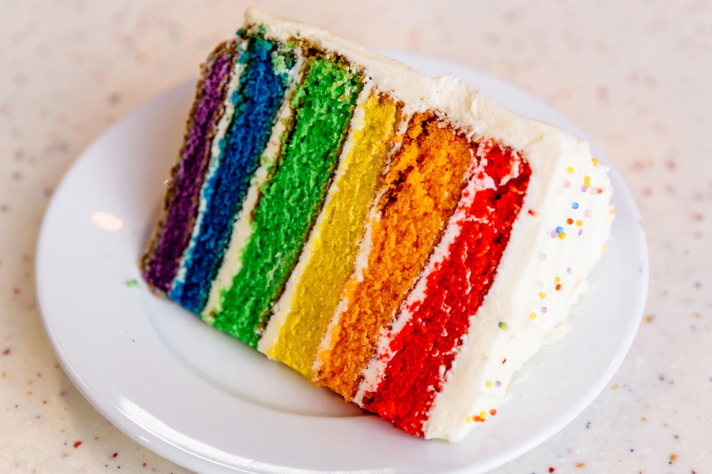 Slice of layer cake with rainbow stripes and white frosting, on a plate with sprinkles on the side