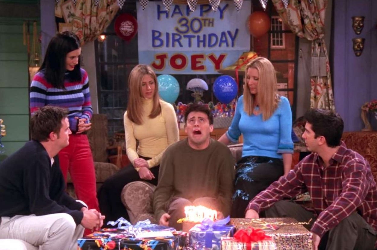 Group of friends celebrate a 30th birthday with a surprised man in the center, surrounded by gifts and a "Happy 30th Birthday Joey" banner