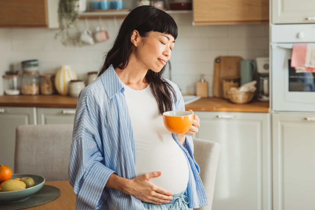 Pregnant person in a kitchen, holding a mug and gently touching their belly, wearing a striped shirt over a plain top
