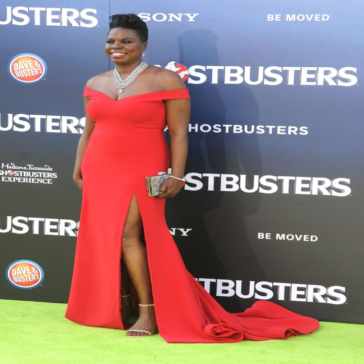 Person in an elegant off-shoulder red gown with a thigh-high slit poses on a red carpet at a "Ghostbusters" event