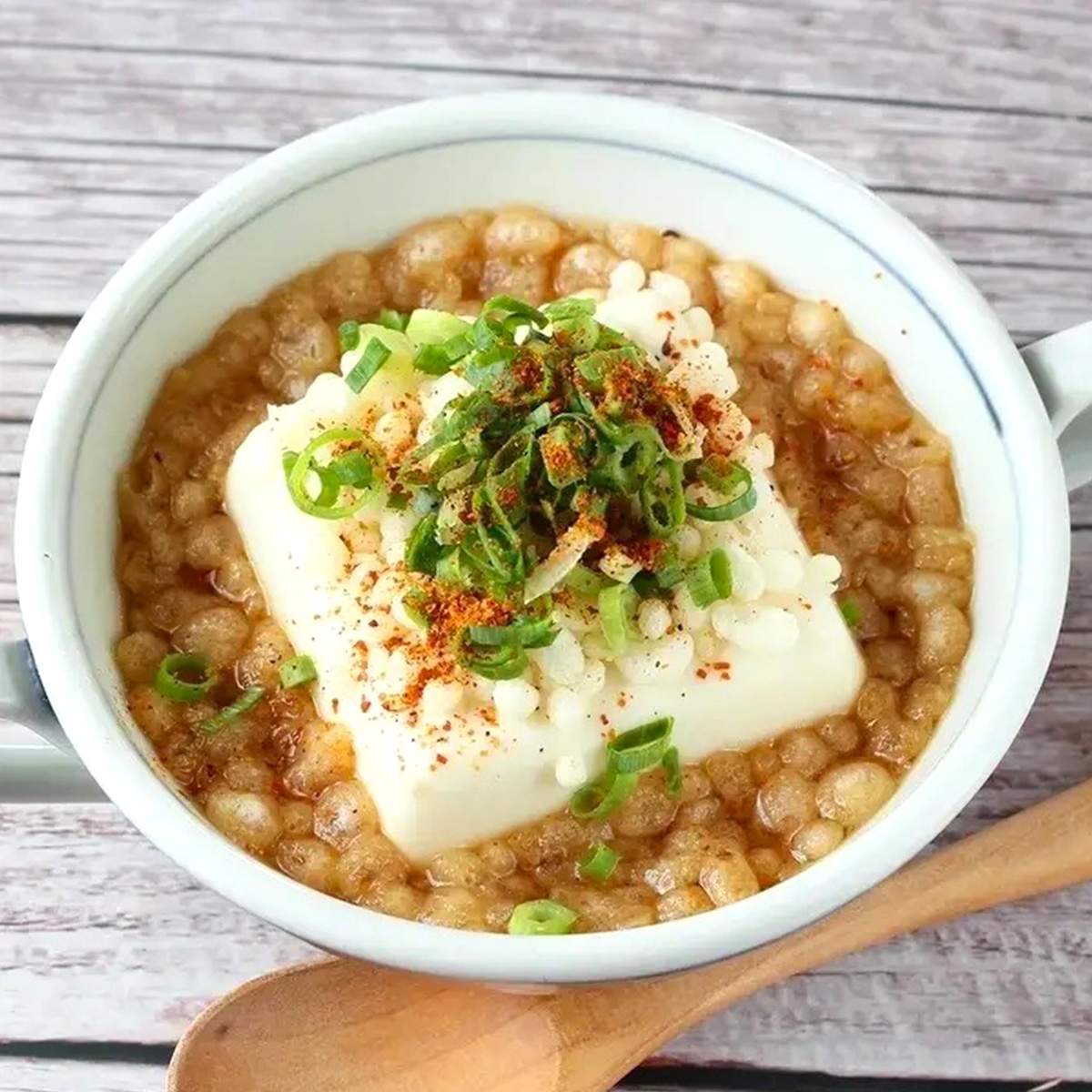 豆腐とネギがトッピングされた温かい麦入りスープが白いボウルに盛られている。木のスプーンが添えられている。