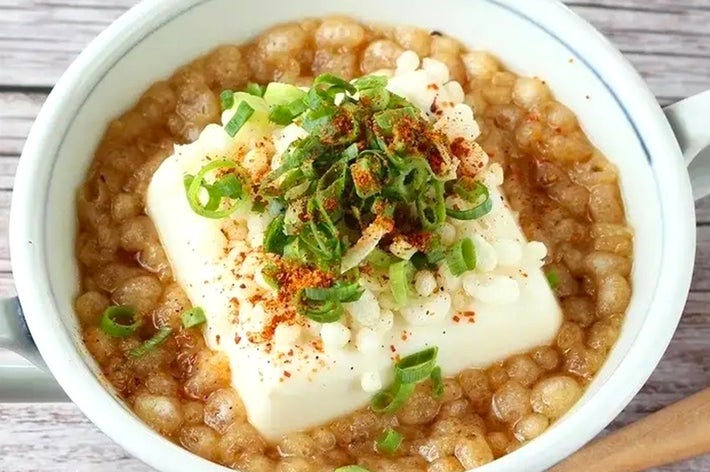 豆腐とネギがトッピングされた温かい麦入りスープが白いボウルに盛られている。木のスプーンが添えられている。