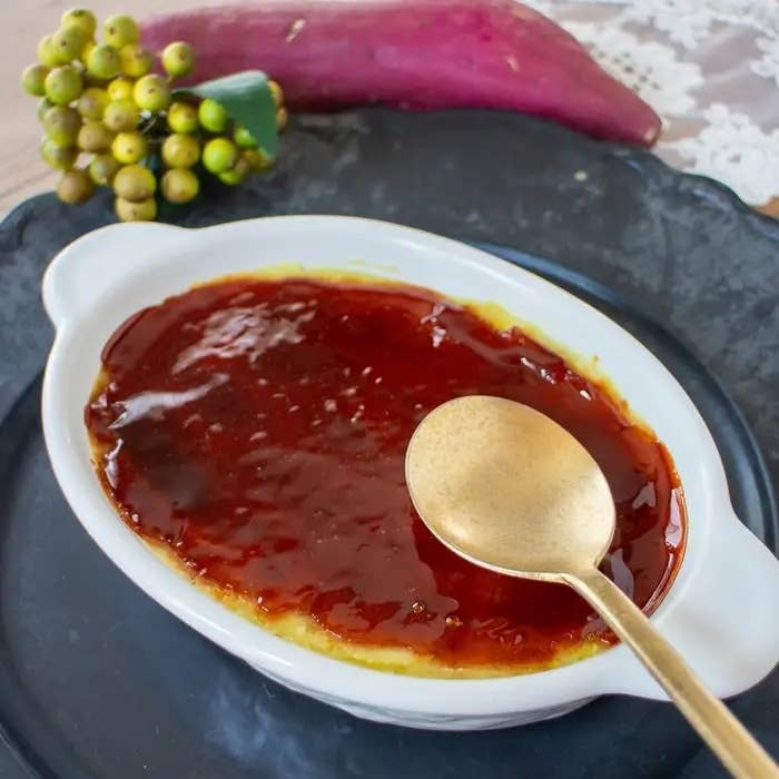 焼き芋のクリームブリュレが皿に盛られ、スプーンが添えられている。背景にはさつまいもとブドウがある。