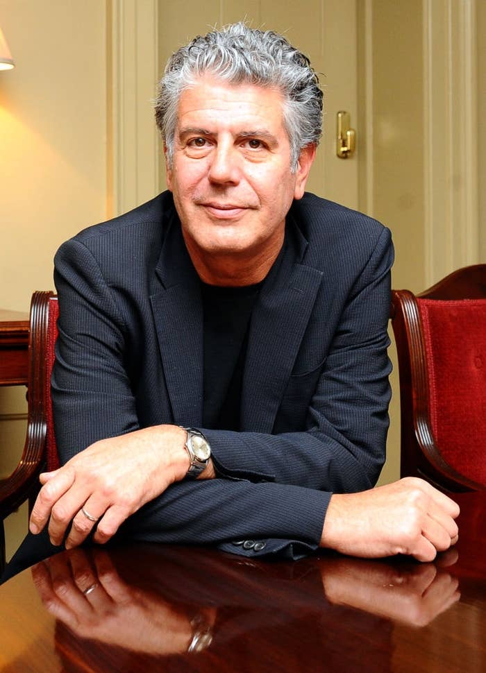 A person with gray curly hair in a dark blazer and black shirt sits at a wooden table, arms crossed