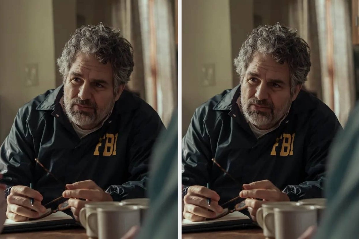 Person with gray hair and beard sits at a table holding glasses, wearing an "FBI" jacket. Appears to be in a thoughtful conversation