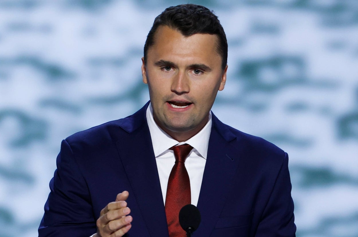A person in a suit and tie speaks passionately at a public event, gesturing with one hand, in front of a blurred background