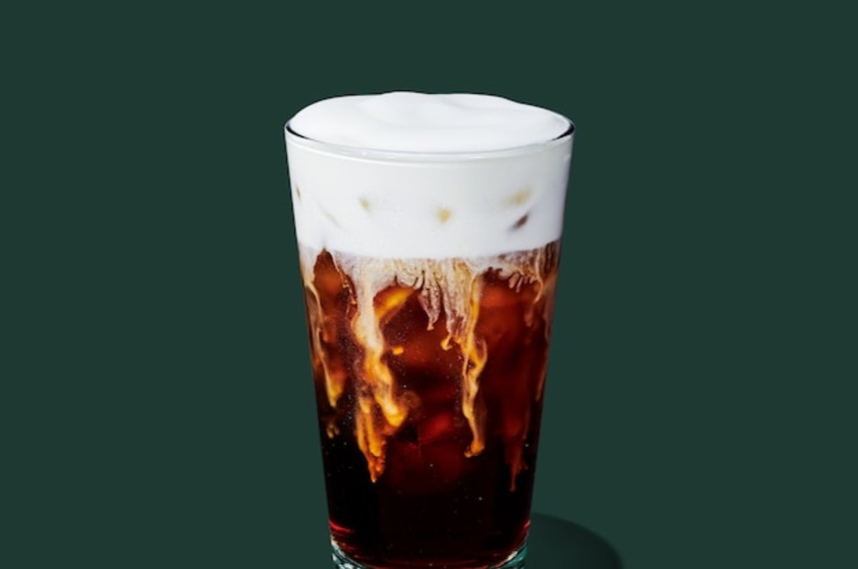 Iced coffee with creamy foam on top in a clear glass, set against a dark background