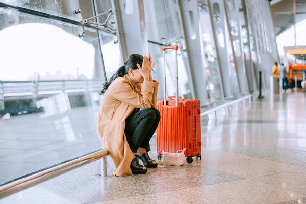 Person sitting on an airport floor in a trench coat, overwhelmed, with a red suitcase and neck pillow beside them