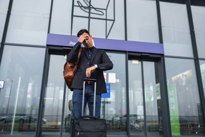Person standing outside an airport, looking stressed with one hand on their forehead and the other on rolling luggage