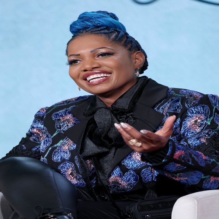 Person smiling, wearing a floral-patterned jacket over a black blouse with shiny leggings, sitting in a casual setting. Hair styled in braids