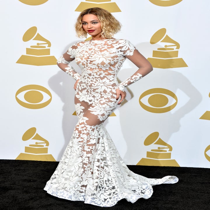 Person in a floor-length, sheer, floral lace gown poses on a music award event red carpet backdrop with trophy graphics