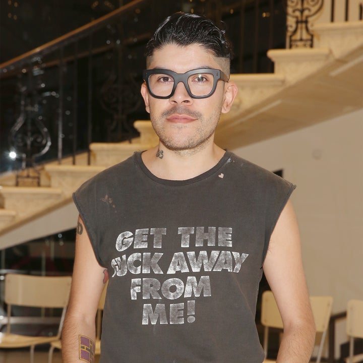 Person with glasses wearing a sleeveless shirt that says "GET THE [CENSORED] AWAY FROM ME!" and black pants, standing indoors