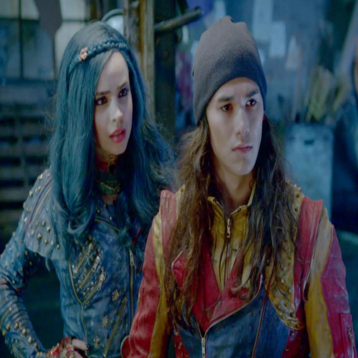 Two people wearing edgy, fantasy-style outfits are in a dimly lit room, looking intently at a third person off-frame