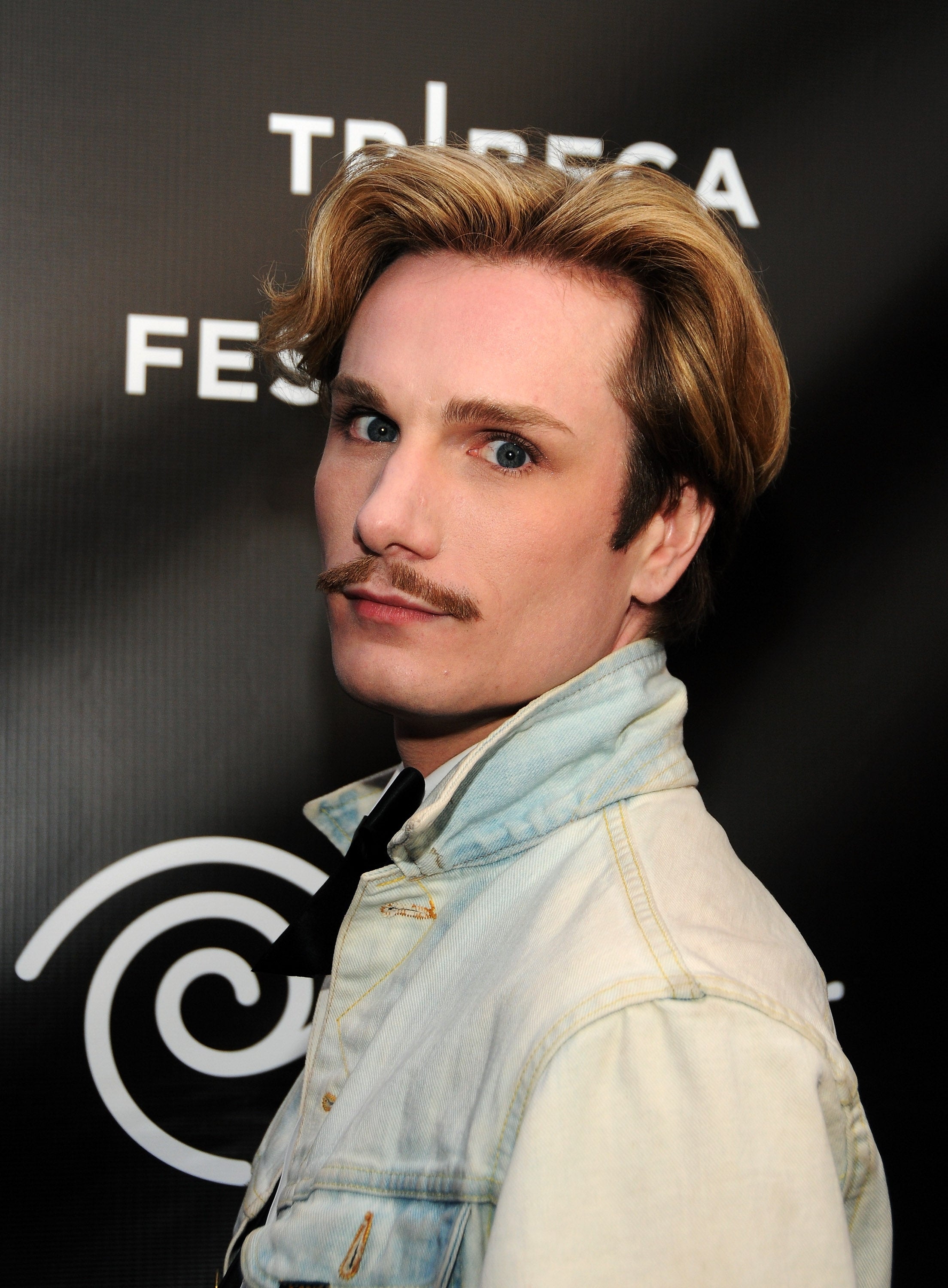 Person with styled hair and mustache wears a denim jacket at Tribeca Festival, posing against a black backdrop with a promotional logo