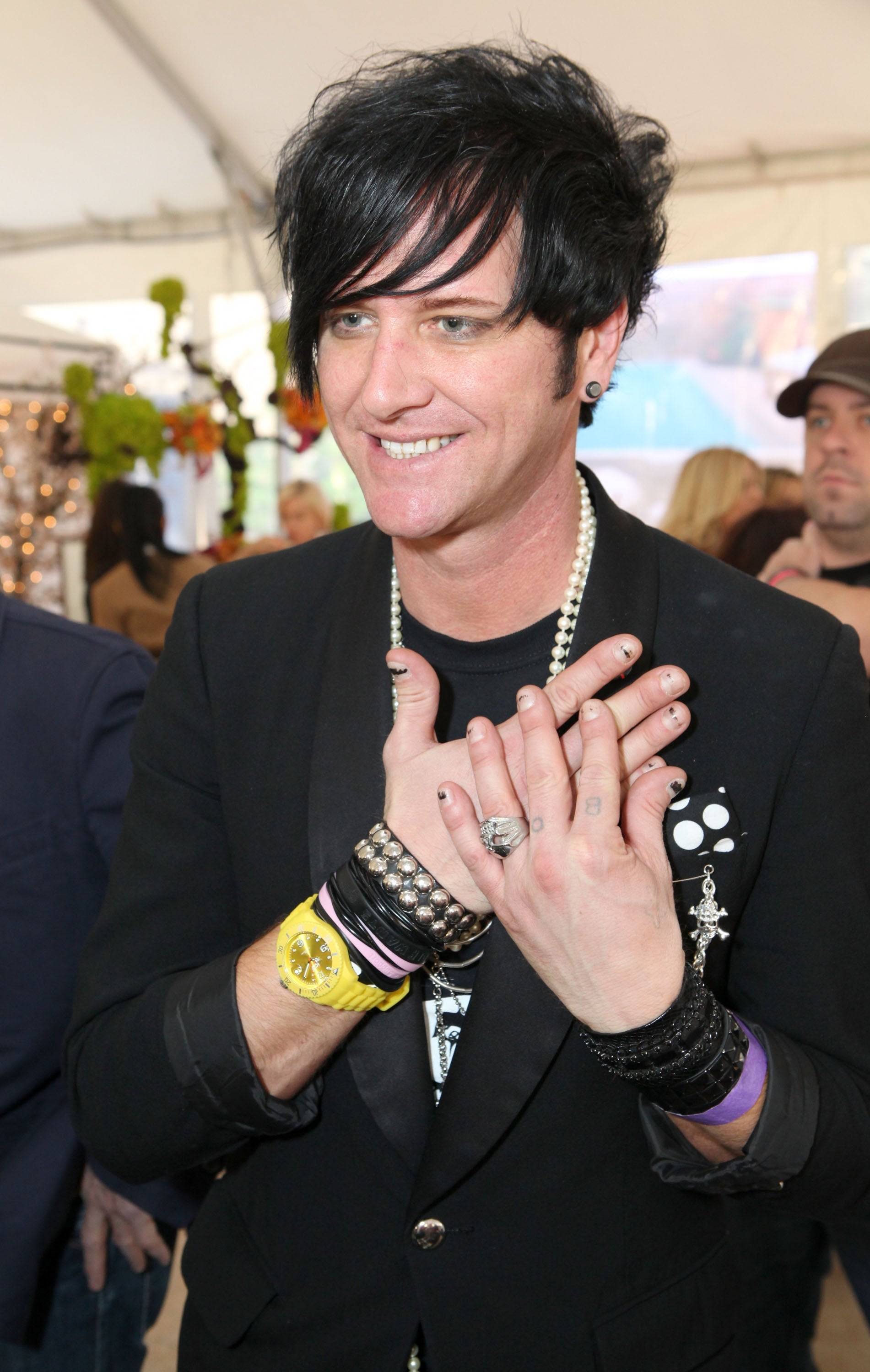 A person in a stylish jacket with multiple bracelets smiles, hands clasped together, at an event