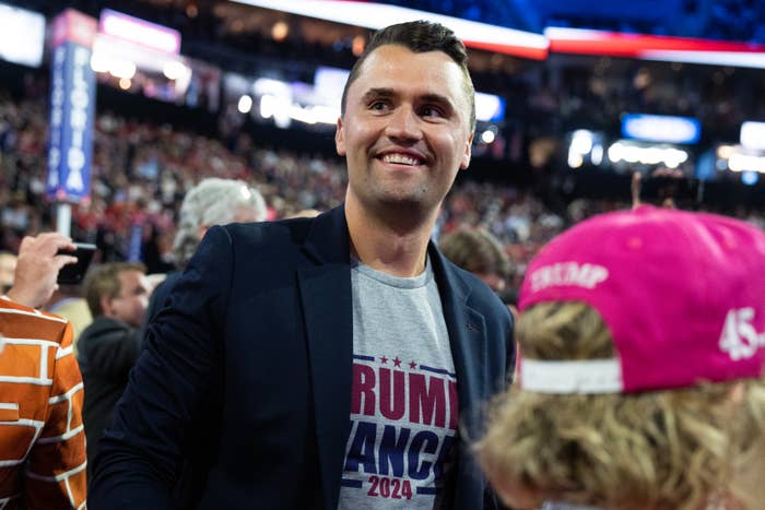 A person in a blazer and campaign t-shirt smiles at a crowded event, with people and signage visible in the background