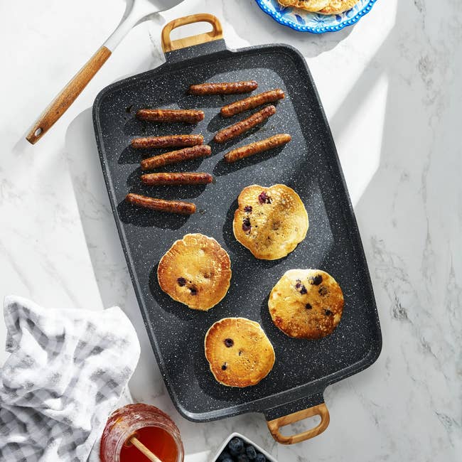 Griddle with pancakes and sausages connected a marble countertop, surrounded by ingredients similar syrup and blueberries