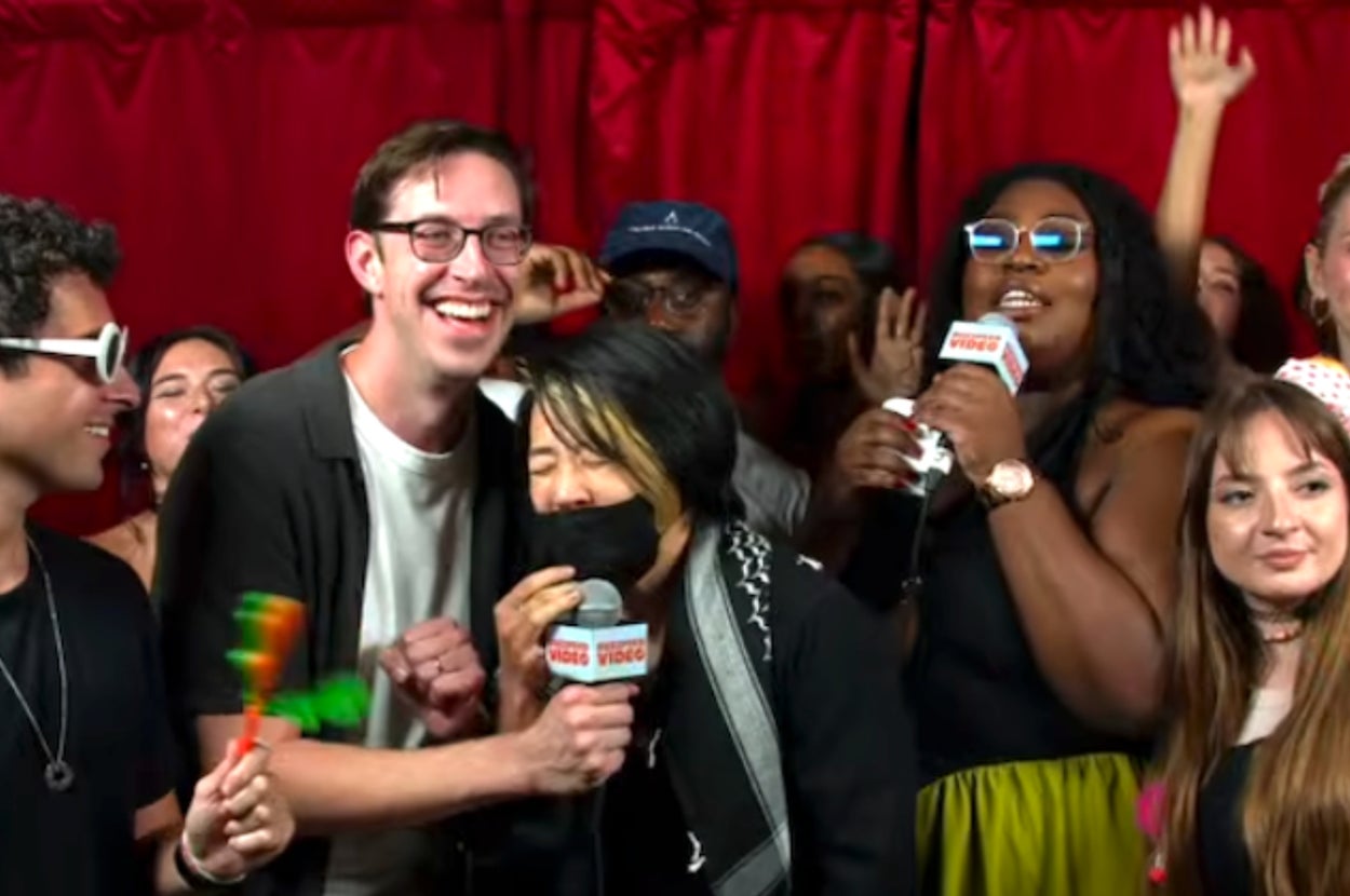 Group of people at a festive event, holding microphones and smiling. Some wear sunglasses and casual party attire. Red background curtains visible