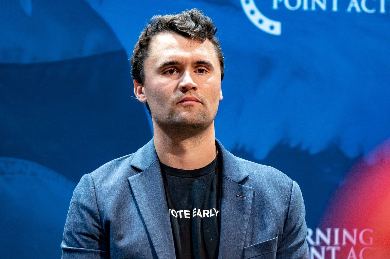 A person in a blazer and t-shirt stands against a blue backdrop at a public event