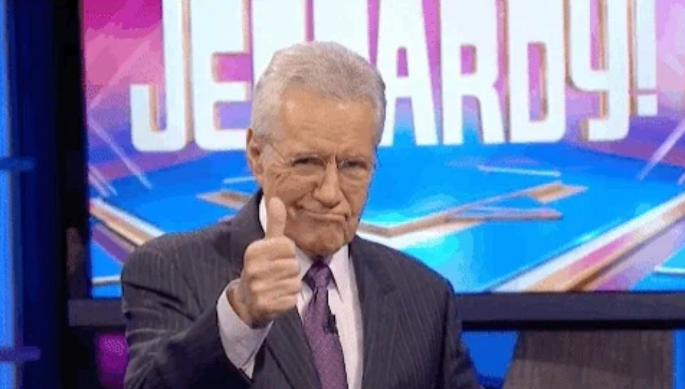 Person giving a thumbs up in front of a "Jeopardy!" sign, wearing a suit and tie