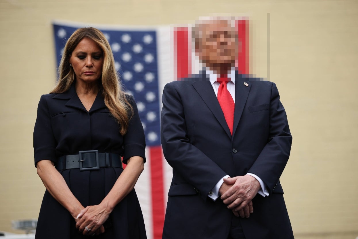 Two individuals stand solemnly in front of a blurred flag. The first person wears a belted, dark dress. The second person wears a suit with a red tie