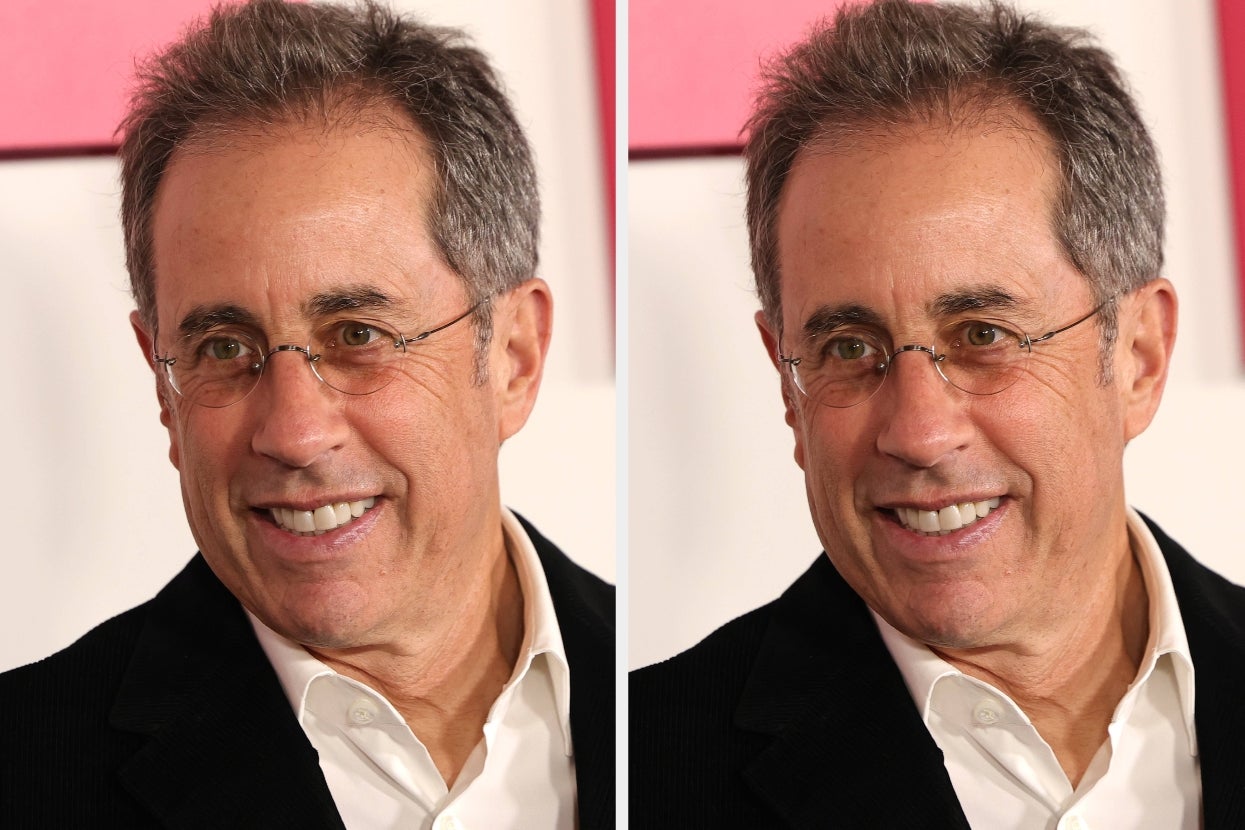 A man with glasses in a suit jacket and open-collared shirt smiles at a red carpet event